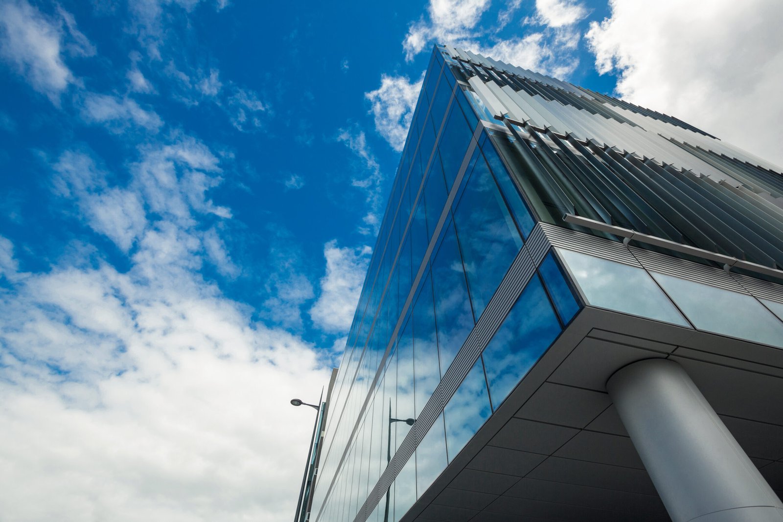 Low-angle view of a glass-clad modern building against a bright blue sky with scattered clouds.